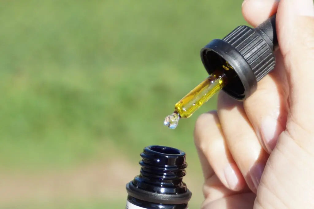 Mão a segurar um conta-gotas com óleo dourado sobre um frasco aberto, com fundo verde desfocado.
