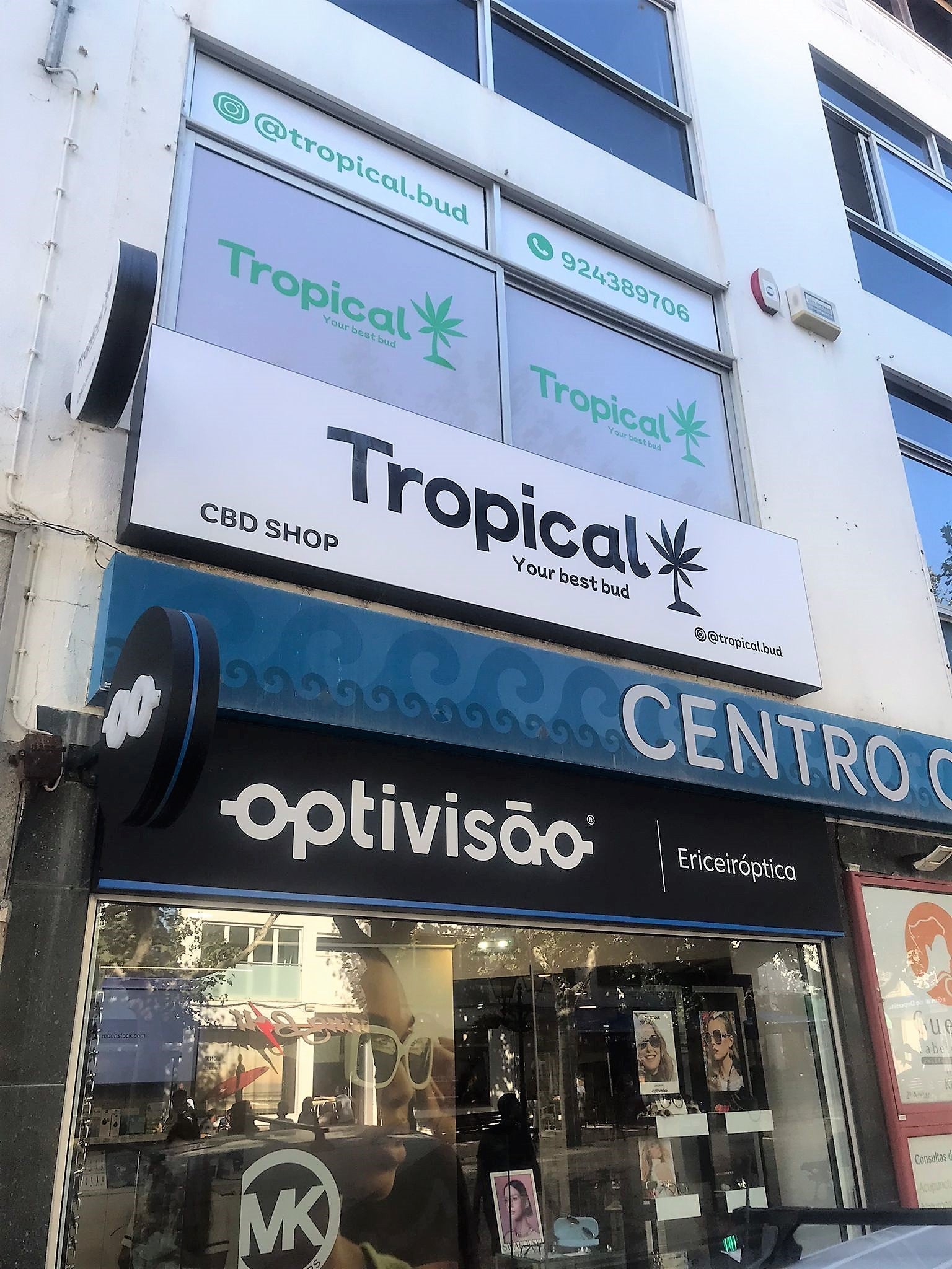 Fachada de um edifício com letreiro e janelas da Tropical CBD Shop na Ericeira, por cima de uma ótica.